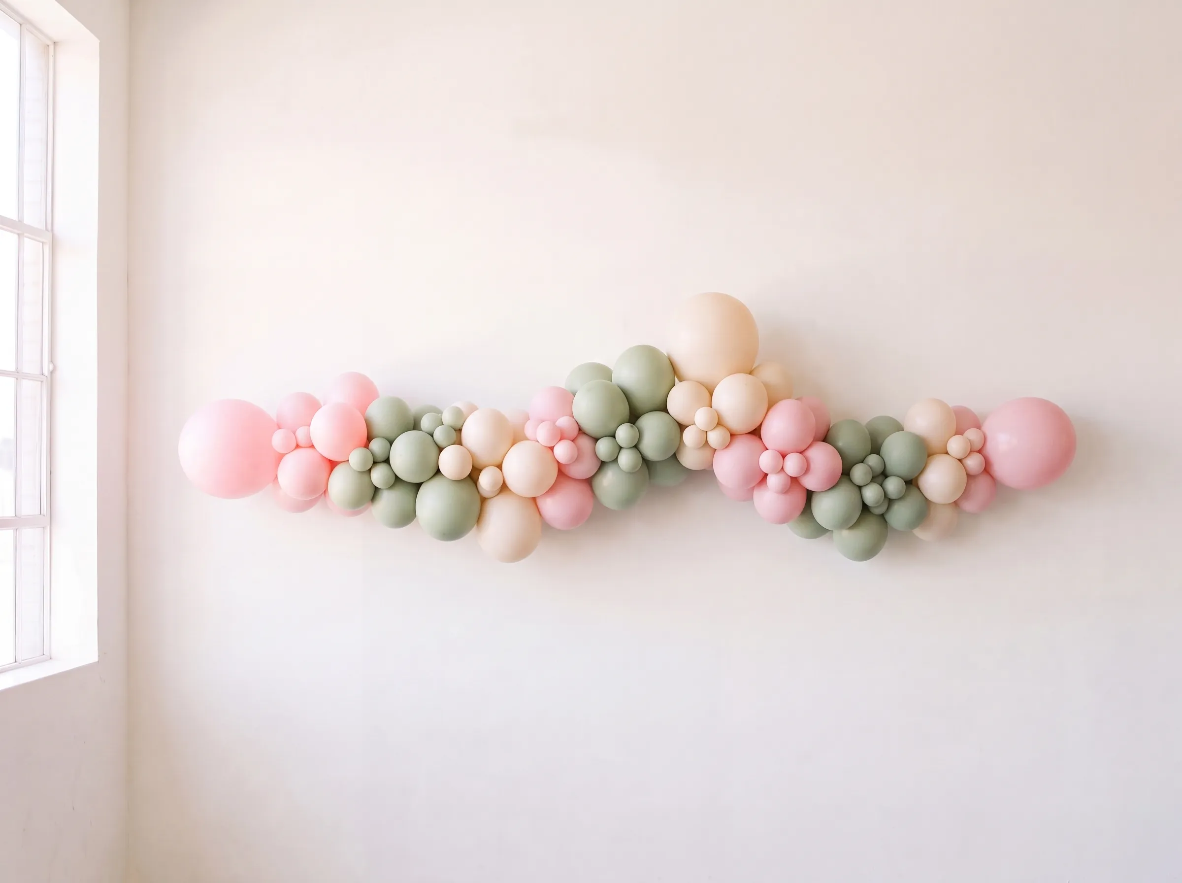 A full organic balloon garland in blush, deep pink and sage against a warm-white wall.