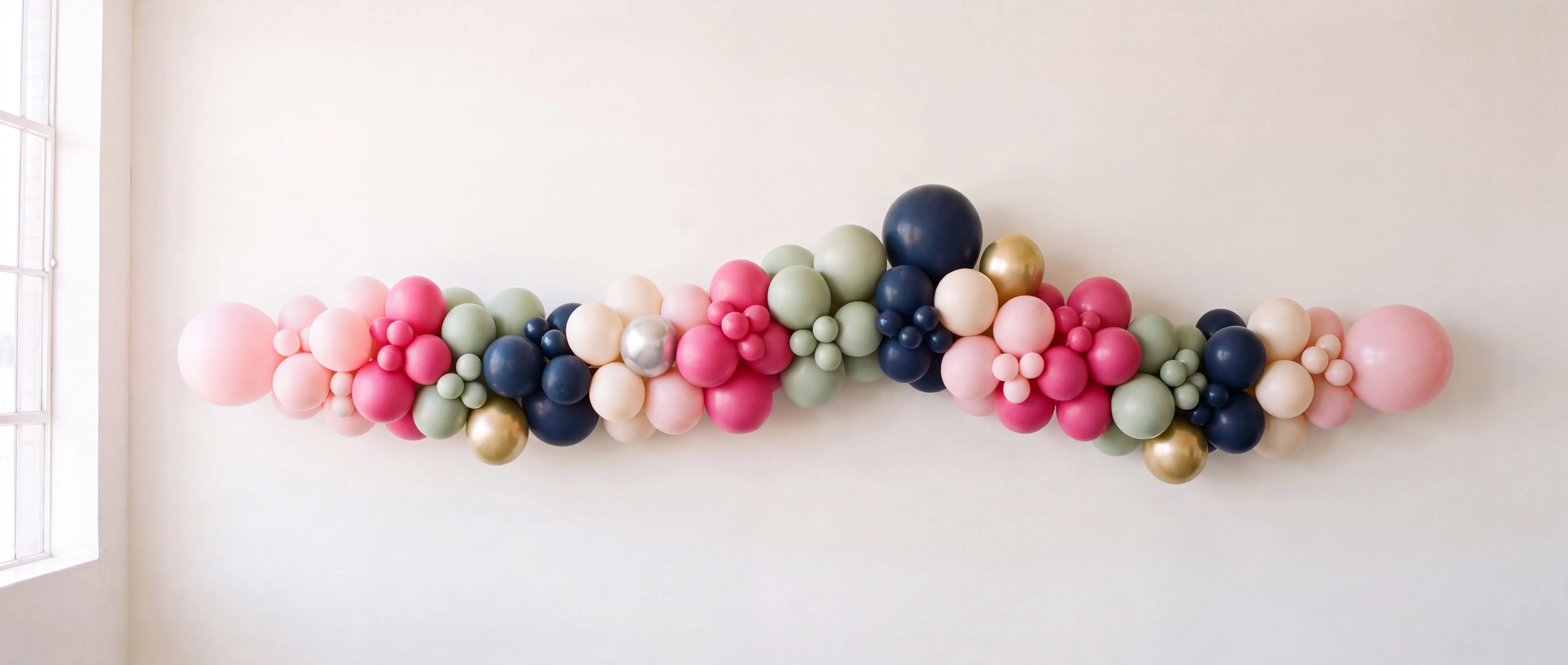 A custom balloon garland in blush, deep pink, sage, navy and gold, installed across a sunlit warm-white wall.